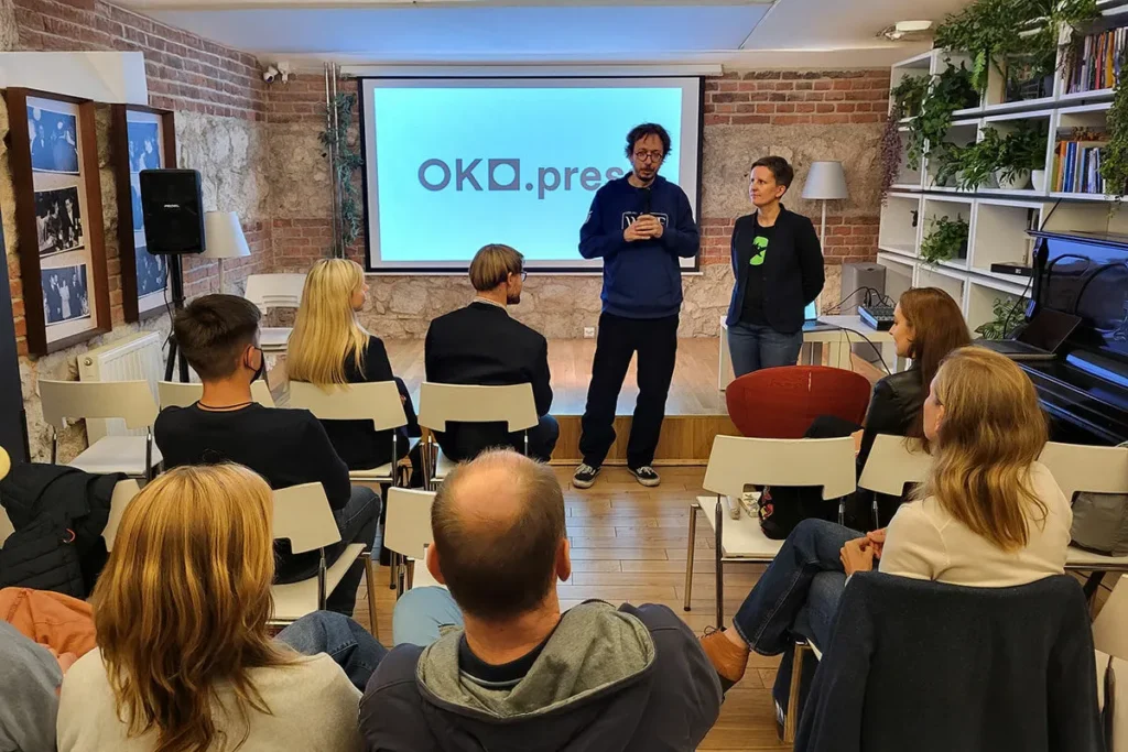 Publiczność siedzi na krzesłach podczas dyskusji w kameralnej sali z ceglaną ścianą. Na scenie dwie osoby prowadzą rozmowę.