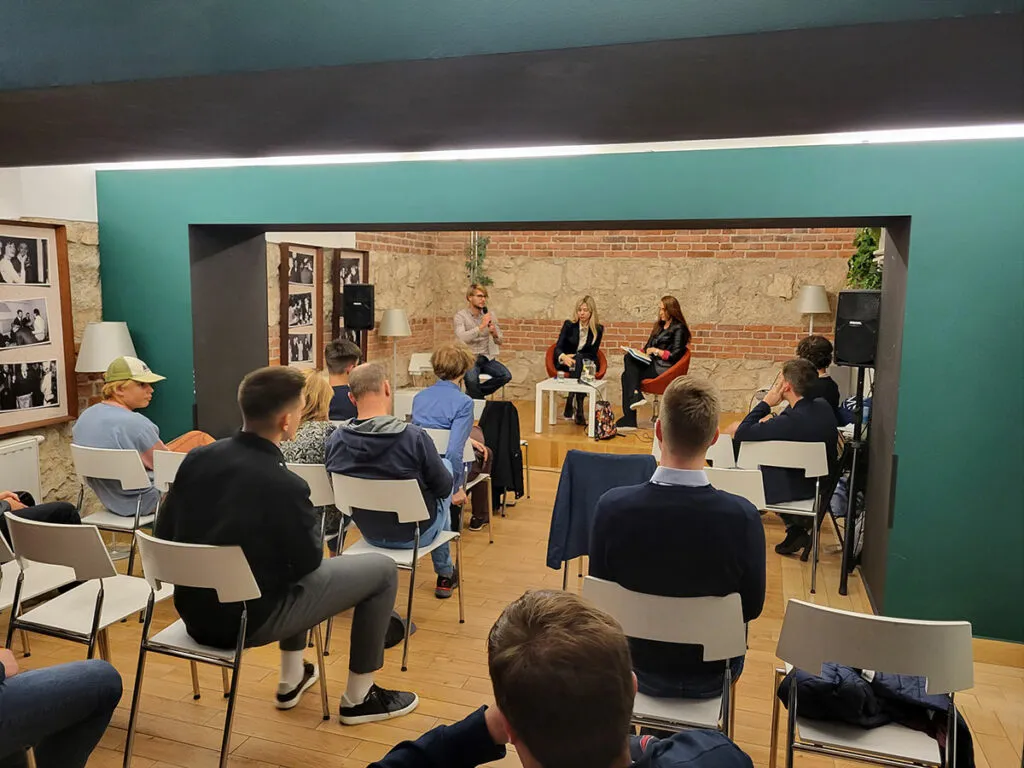 Uczestnicy spotkania siedzą na krzesłach, słuchając rozmowy trzech prelegentów na scenie. Kobieta z mikrofonem gestykuluje, a obok niej siedzą pozostali rozmówcy. Wnętrze ma ceglane ściany i drewnianą podłogę.