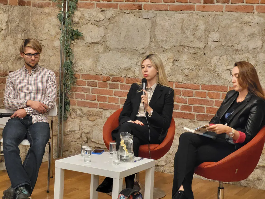 Trzy osoby siedzą na scenie podczas panelu dyskusyjnego. Kobieta w środku mówi do mikrofonu, obok niej siedzi mężczyzna oraz druga kobieta z książką w ręku. W tle ceglana ściana i zieleń.