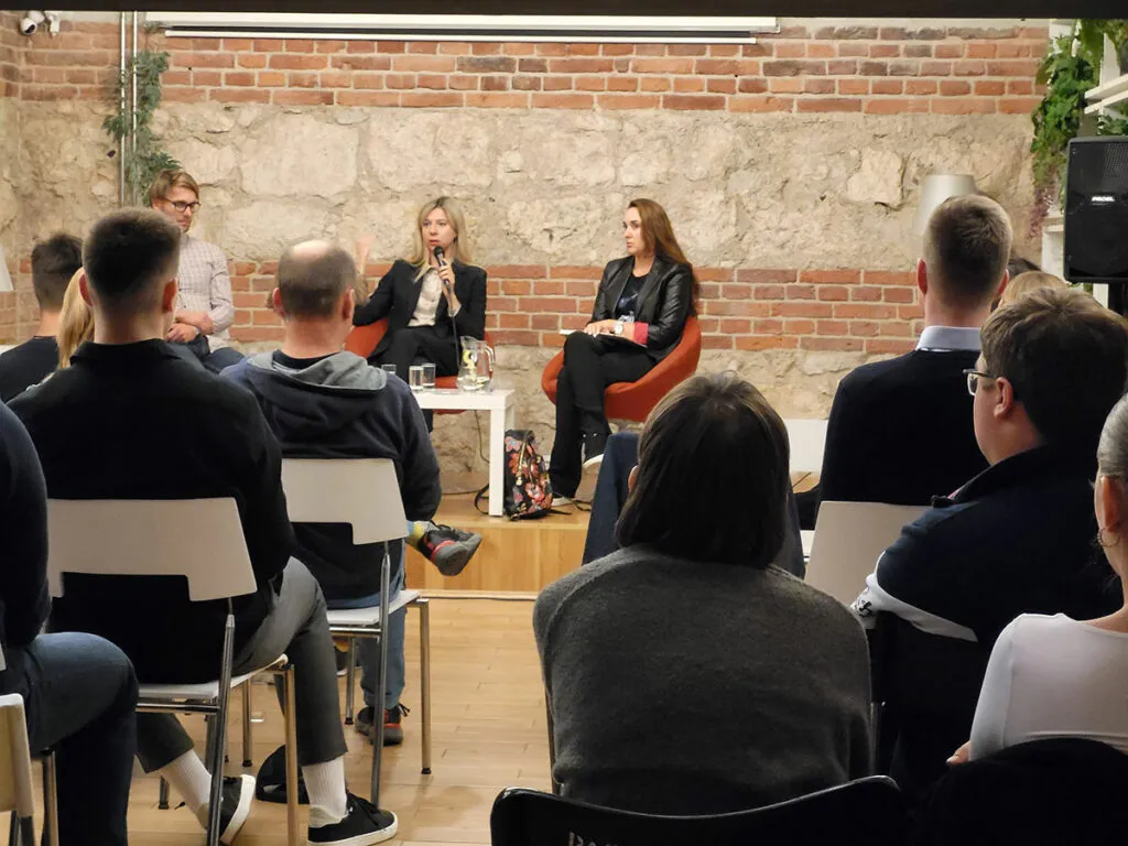 Publiczność siedzi na krzesłach podczas dyskusji w kameralnej sali z ceglaną ścianą. Na scenie trzy osoby prowadzą rozmowę przy stoliku z mikrofonami.