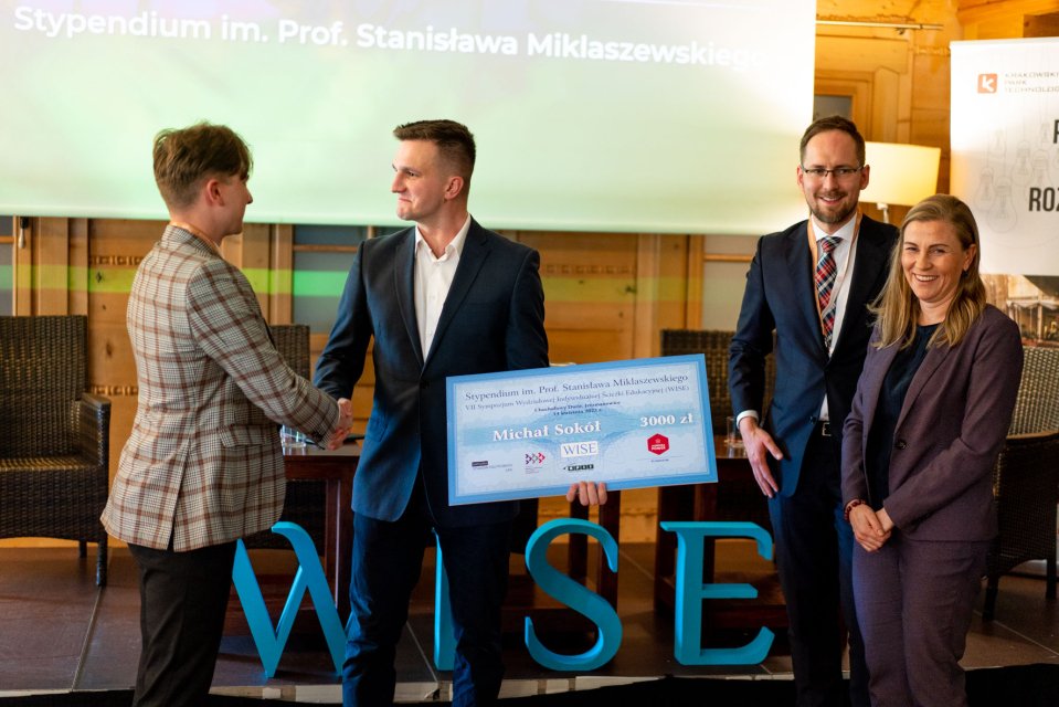 Z lewej strony 2 mężczyzn ściskających dłonie, jeden z nich trzyma duży czek. Z prawej mężczyzna i kobieta.